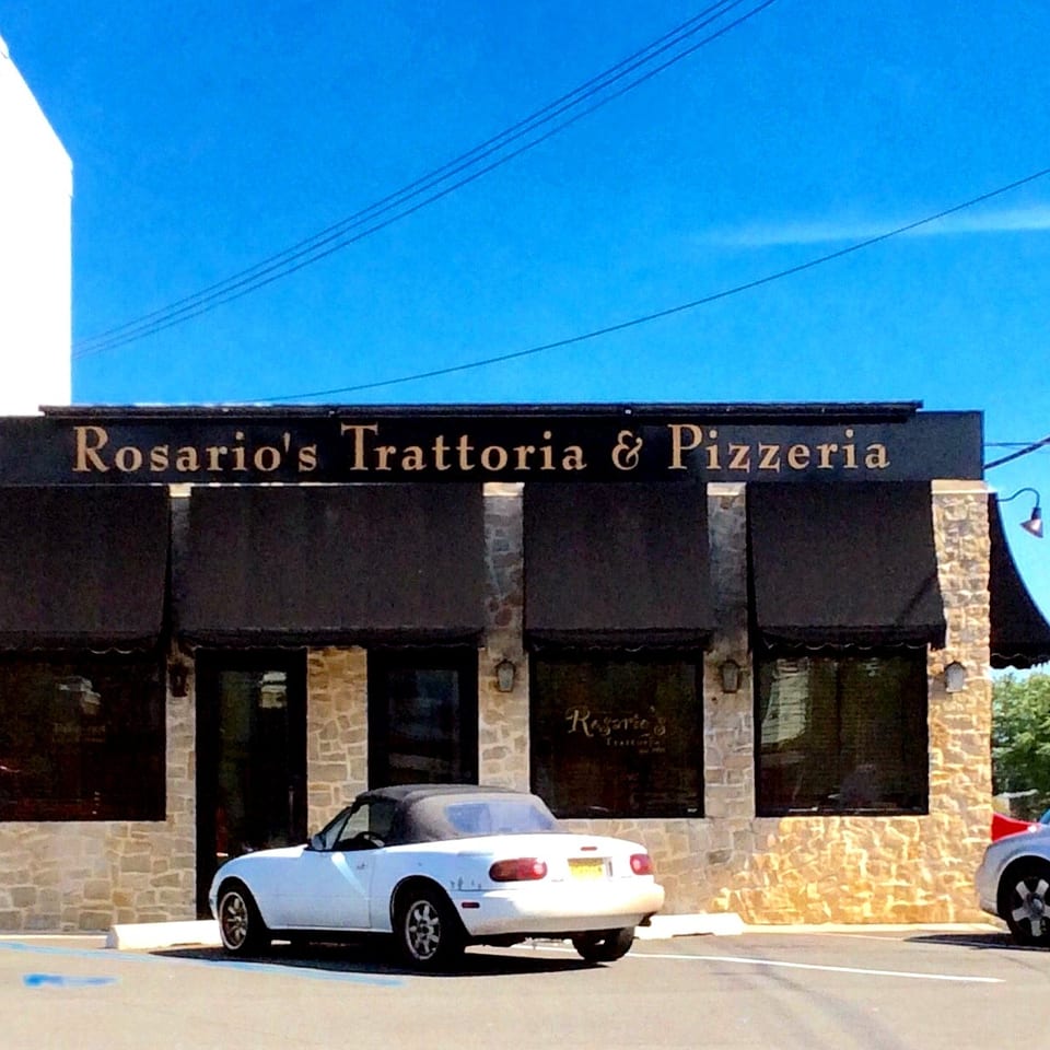 Rosario's Trattoria & Pizzeria storefront with stone facade, black awnings, parked cars, and bright blue sky.