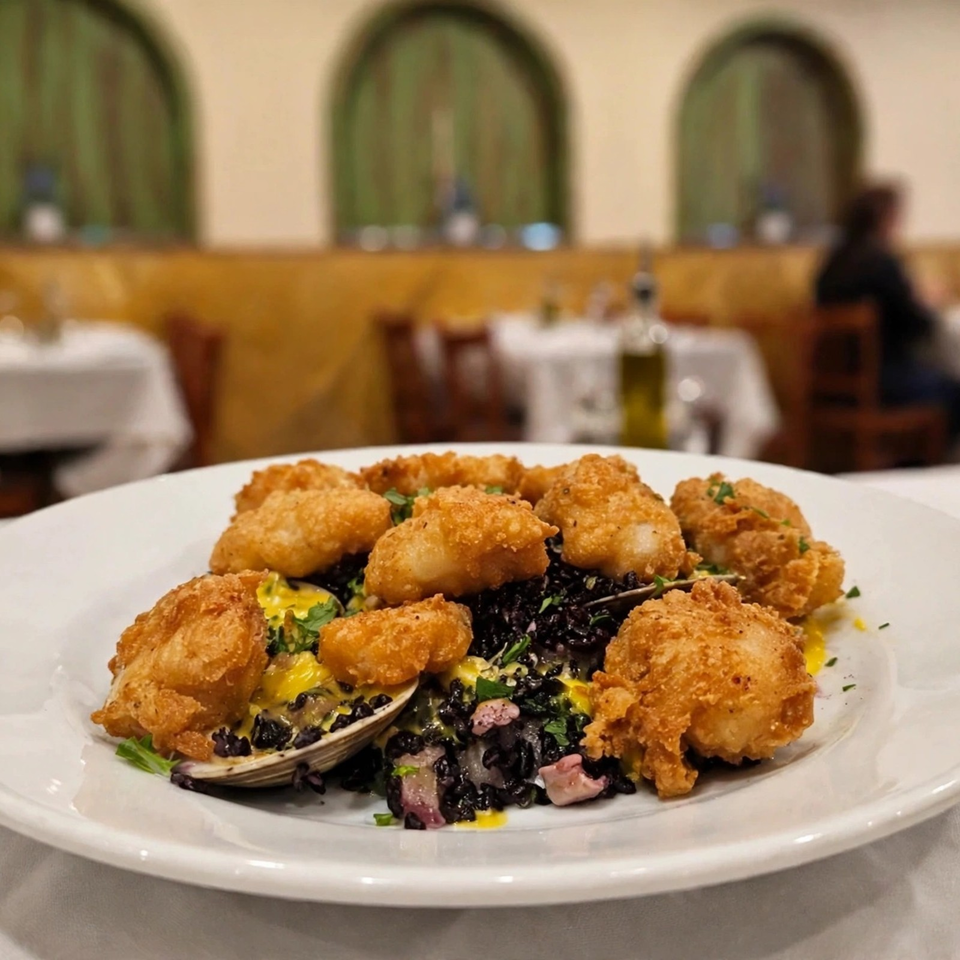 Plate of golden fried seafood over black rice with clams, greens, and sauce in an elegant restaurant dining room.