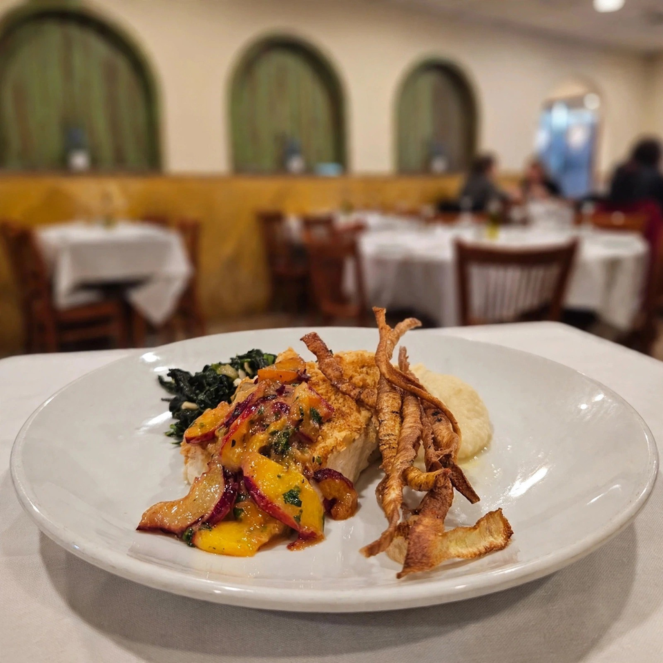 Plated entrée with sauced fish, roasted vegetables, greens, crispy root vegetables, and puree in an elegant restaurant dining room.