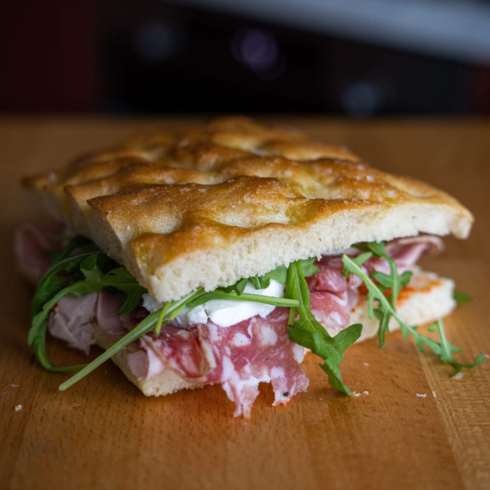 Italian focaccia sandwich with salami, prosciutto, fresh mozzarella, and arugula on a wooden surface.