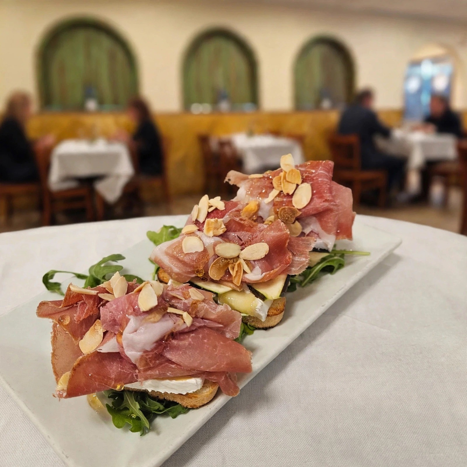 Plate of toasted bread topped with prosciutto, greens, cheese, honey, and sliced almonds in an elegant restaurant dining room.