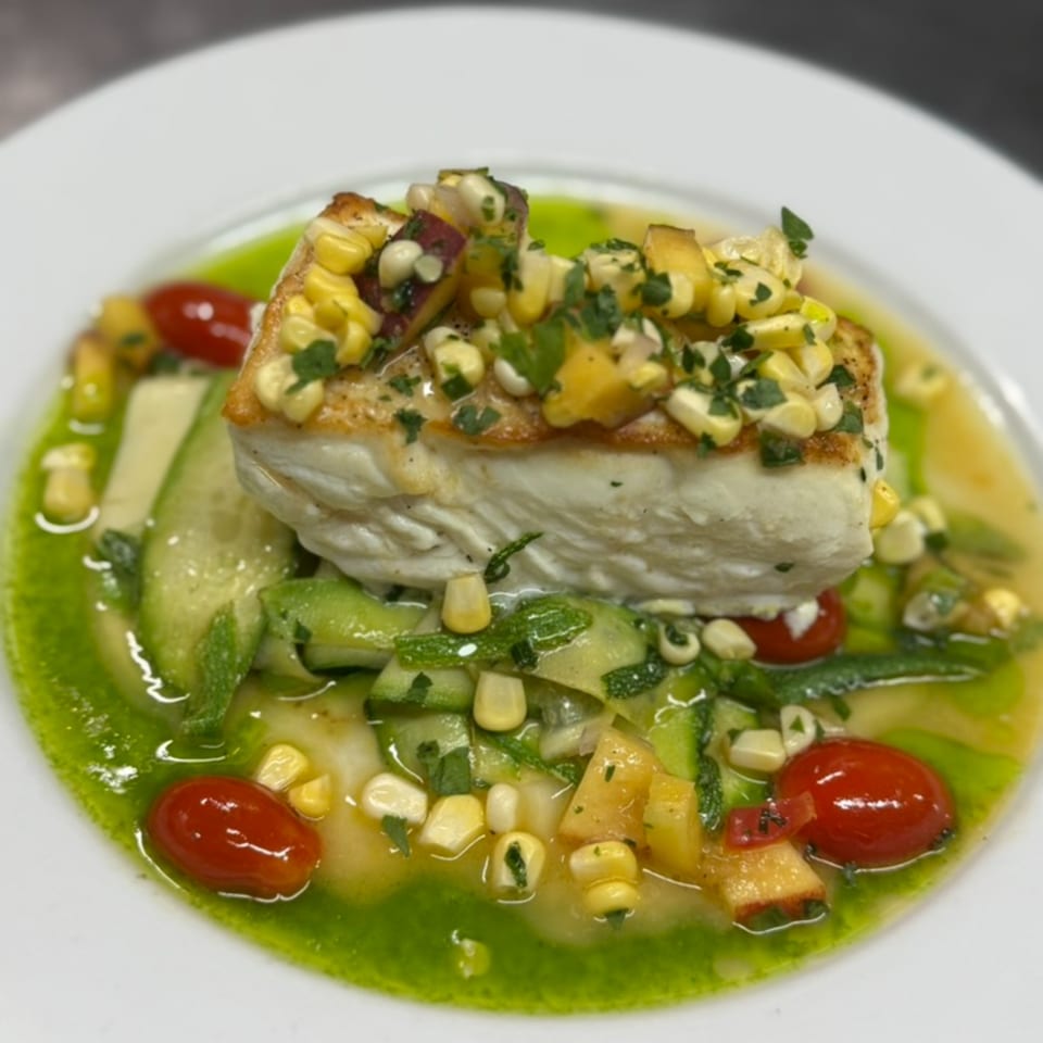 Pan-seared fish with corn and cherry tomatoes, drizzled with green sauce, served on a white plate.