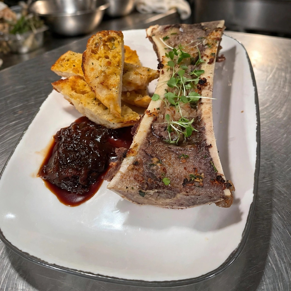 Roasted bone marrow topped with microgreens, garlic bread wedges, and dark dipping sauce on a white plate.