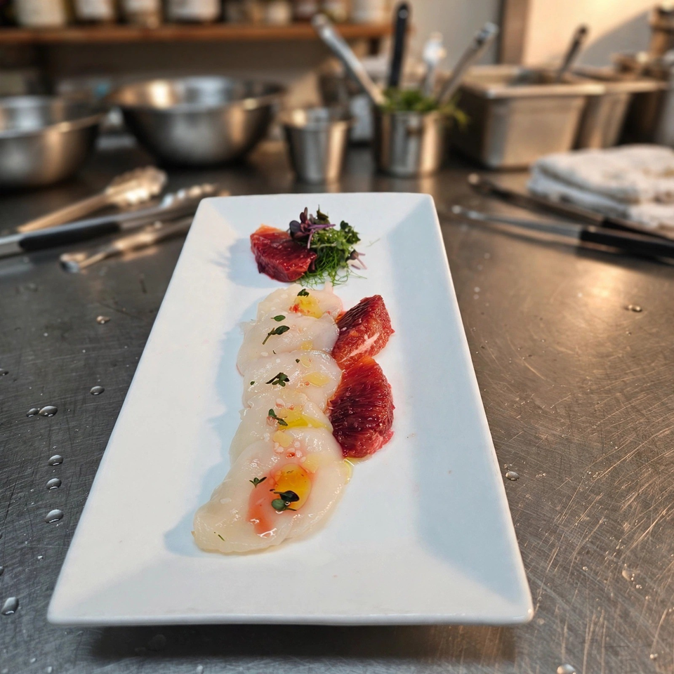 Plated scallops with blood orange segments, microgreens, and citrus garnish on a white rectangular plate in a professional kitchen.