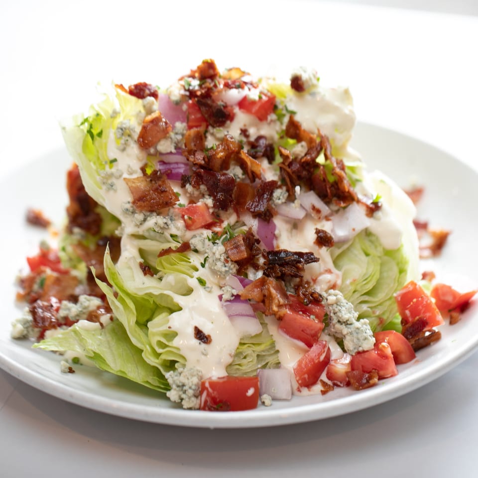 A wedge salad with blue cheese dressing, bacon, tomatoes, and red onion on a white plate.