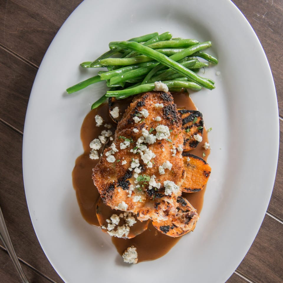Breaded chicken with blue cheese, grilled sweet potatoes, green beans, and brown sauce on a white plate.