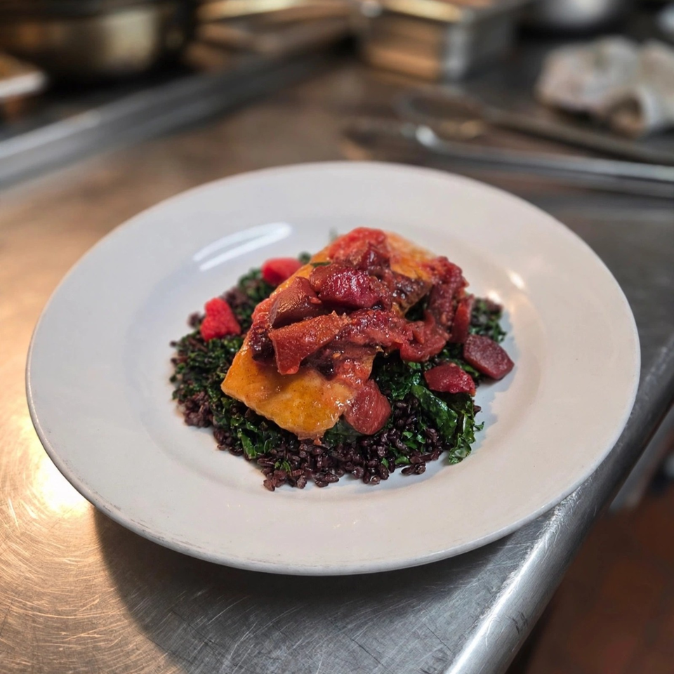 Plated entrée featuring roasted meat, red fruit sauce, leafy greens, and black rice on a white plate in a restaurant kitchen.