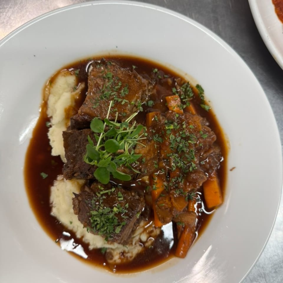 Braised short ribs with mashed potatoes, carrots, and gravy, garnished with fresh herbs.