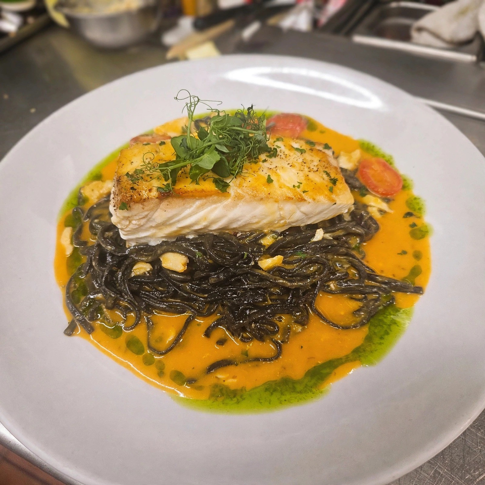 Seared white fish fillet atop black noodles, orange cream sauce, cherry tomatoes, herbs, and green oil on a white plate.