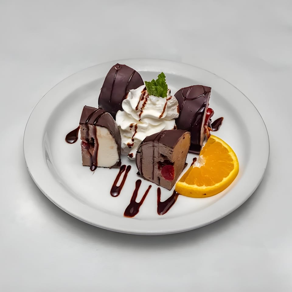 Chocolate-covered dessert pieces with whipped cream, mint, chocolate drizzle, and an orange slice on a white plate.