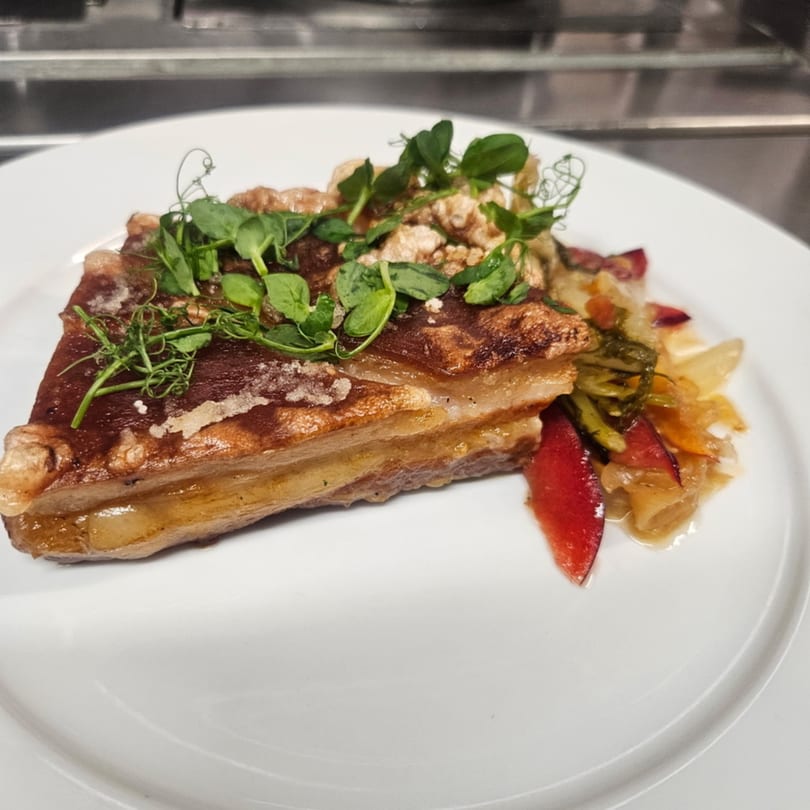 Crispy pork belly with pea shoots and a side of sautéed vegetables on a white plate.