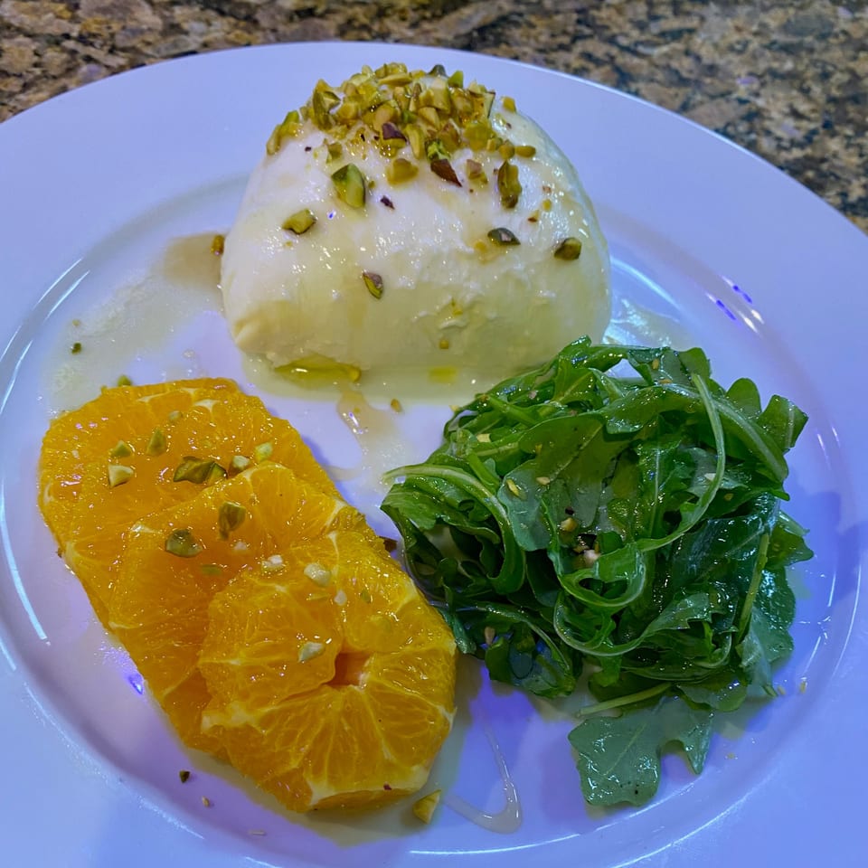 Burrata cheese topped with pistachios, served with orange slices and arugula on a white plate.